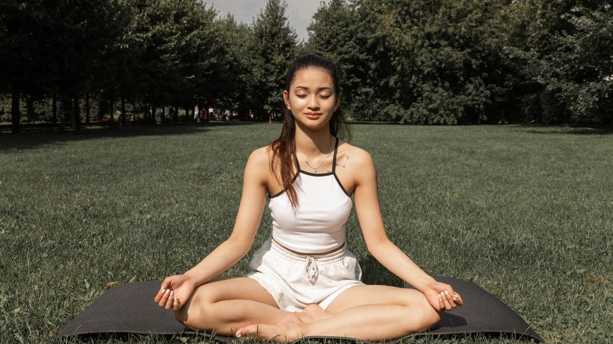 Full length of young Asian female in activewear sitting on mat while meditating in sunny park