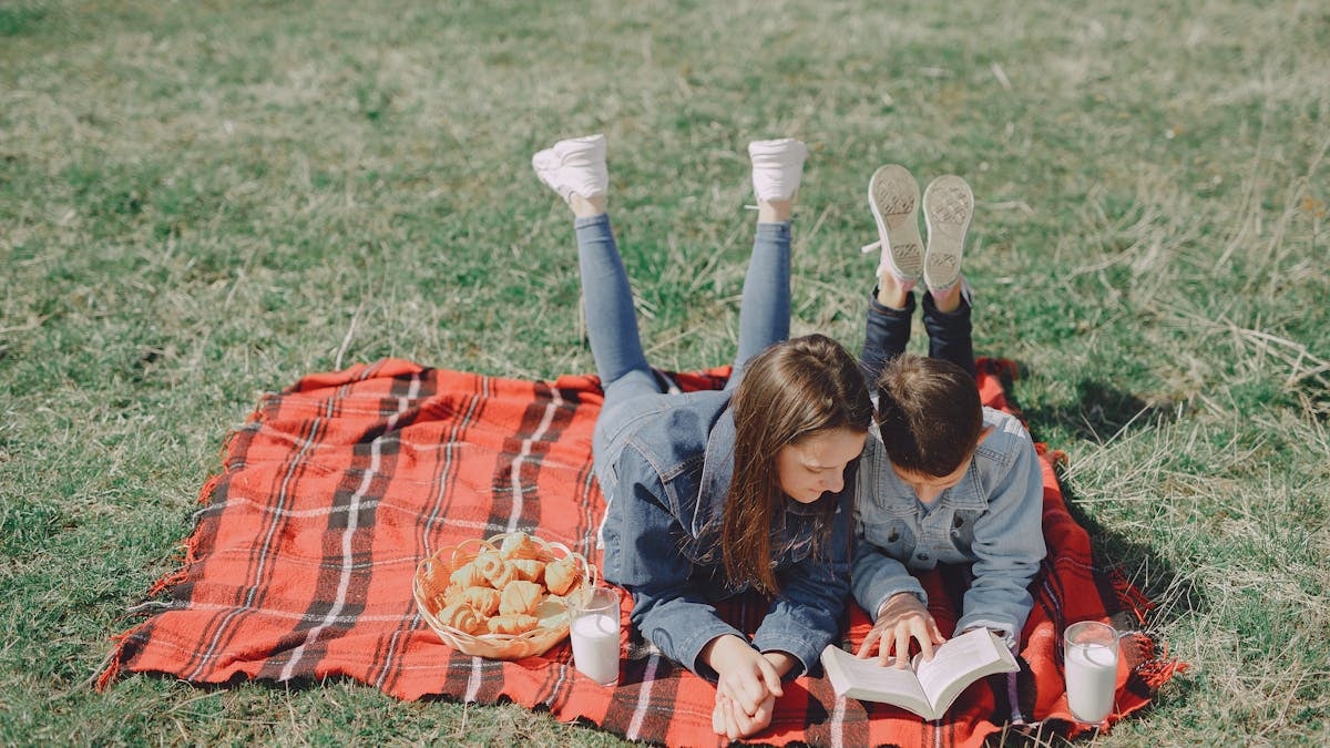 From above of boy and girl in trendy clothes lying together on checkered blanket and reading book while enjoying picnic in countryside during holiday